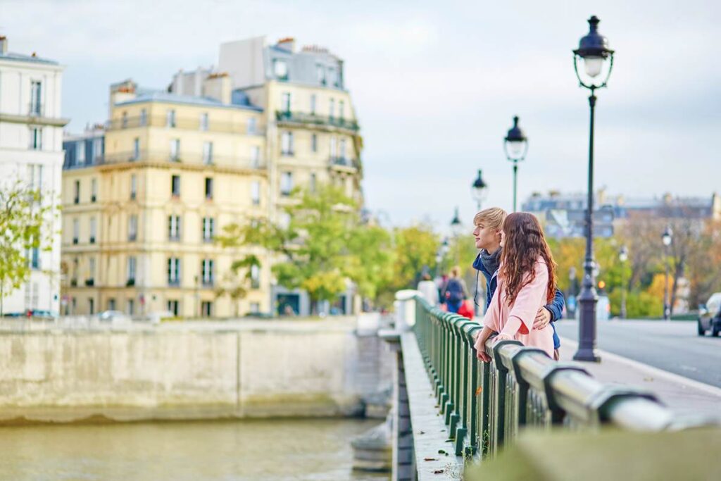A couple enjoys a scenic view on a Paris bridge, with historic buildings and street lamps in the background.