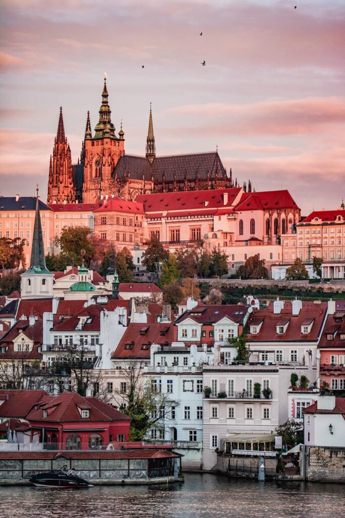 View of Prague Castle with sunset sky, red-roofed buildings, and river reflections in Prague, Czech Republic.