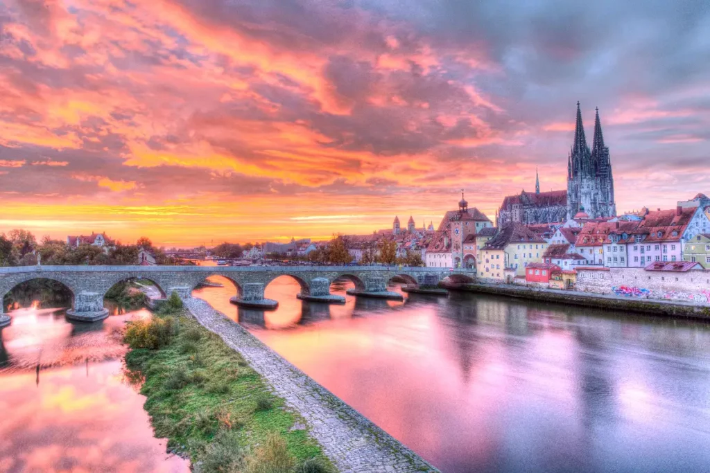 Sunset over a river in Regensburg, Germany, with a historic bridge and colorful houses illuminated by the sky.