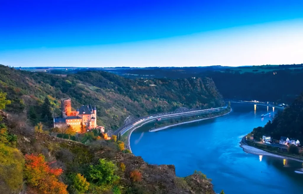 Scenic view of a medieval castle by the Rhine River, surrounded by lush hills and a vibrant autumn landscape.