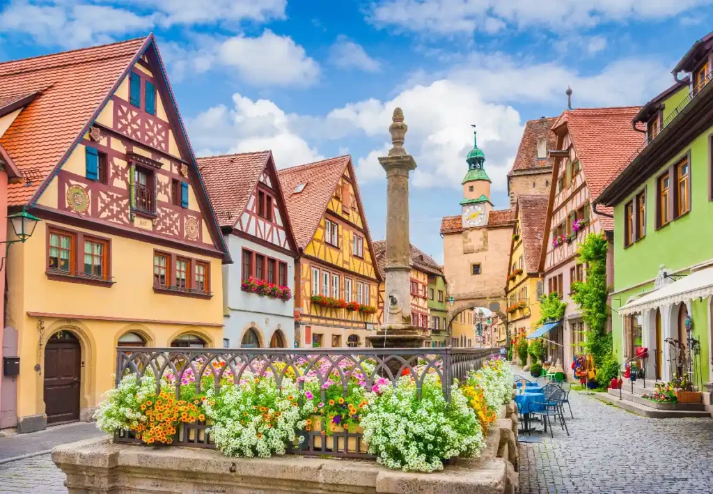 Charming European village with colorful half-timbered houses, flowers, and a historical fountain under a blue sky.