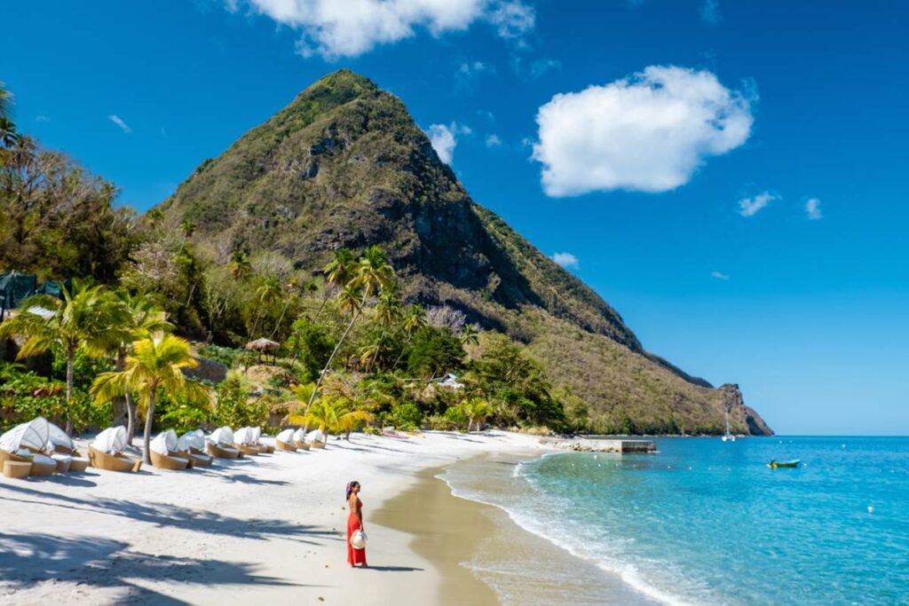 Tropical beach with palm trees, mountain, and blue sea under a sunny sky. Woman in red dress walking on sand.