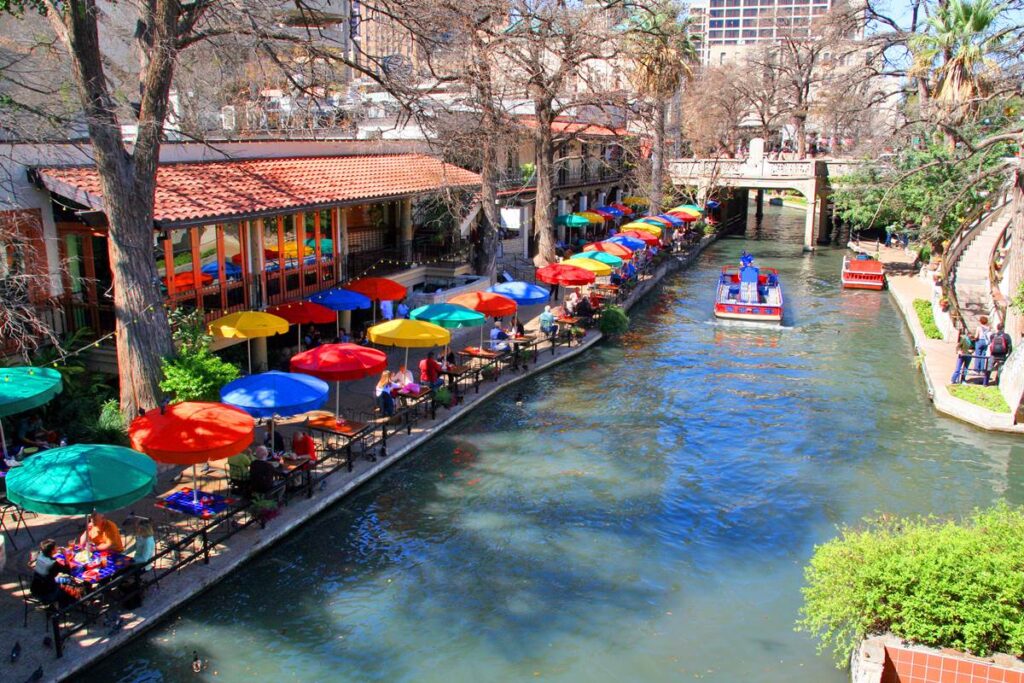 Colorful umbrellas line the San Antonio River Walk, with tourists enjoying dining and views along the water.