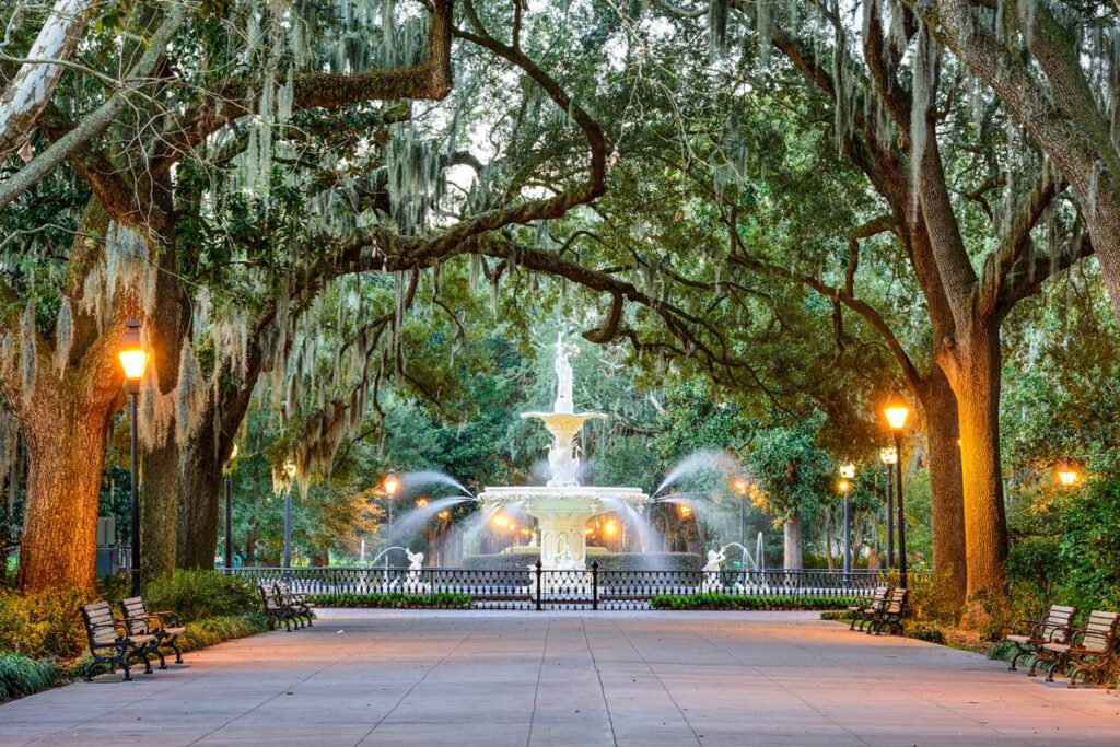 Beautiful fountain in Forsyth Park, Savannah, beneath moss-covered oak trees, surrounded by benches and warm lighting.