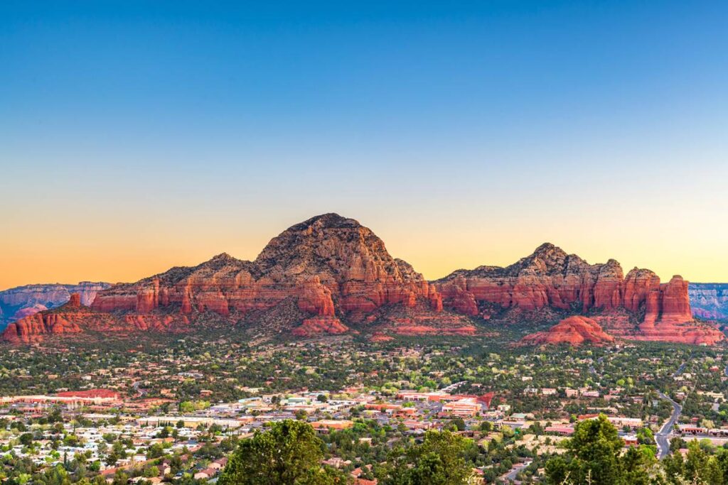 Sunset over Sedona, Arizona, highlighting vibrant red rock formations and the sprawling town below, under a clear sky.