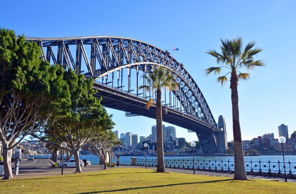 Sydney Harbour Bridge over sparkling water with a clear blue sky and surrounding trees and skyline.