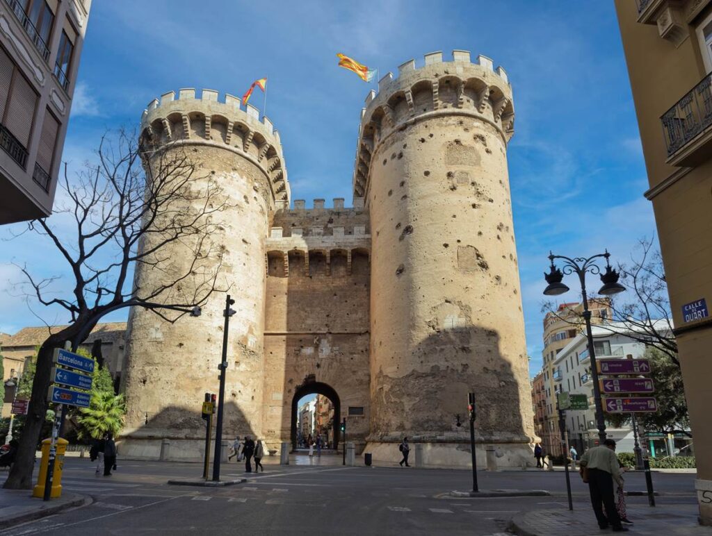 Two ancient stone towers in Valencia, Spain, under a clear blue sky with people walking nearby.