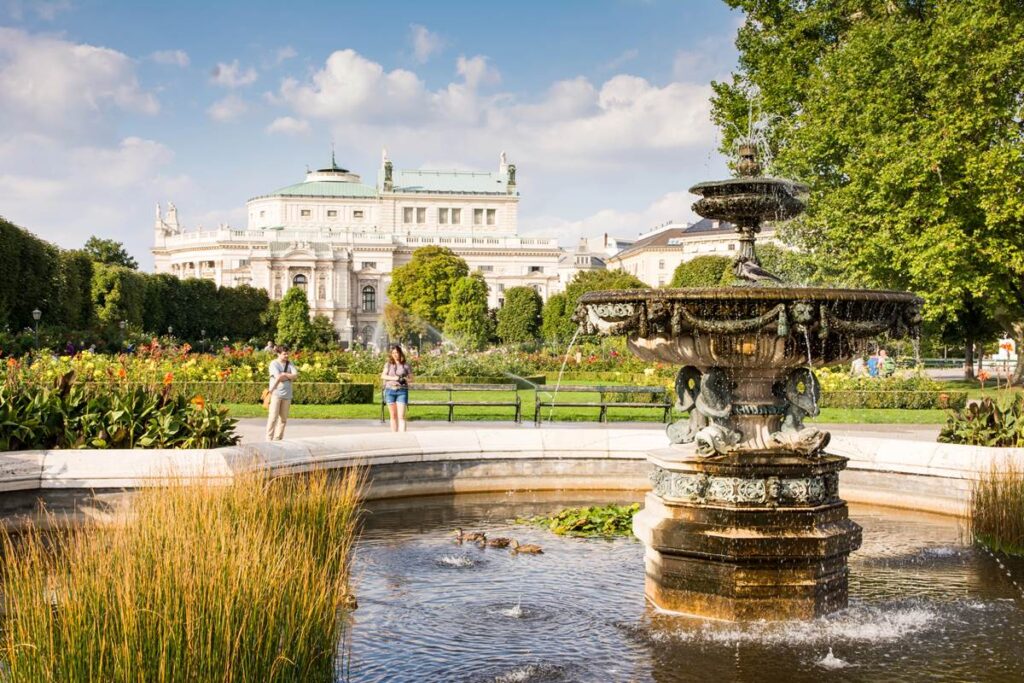 Elegant fountain in a lush garden with a historic building in the background under a clear, sunny sky.