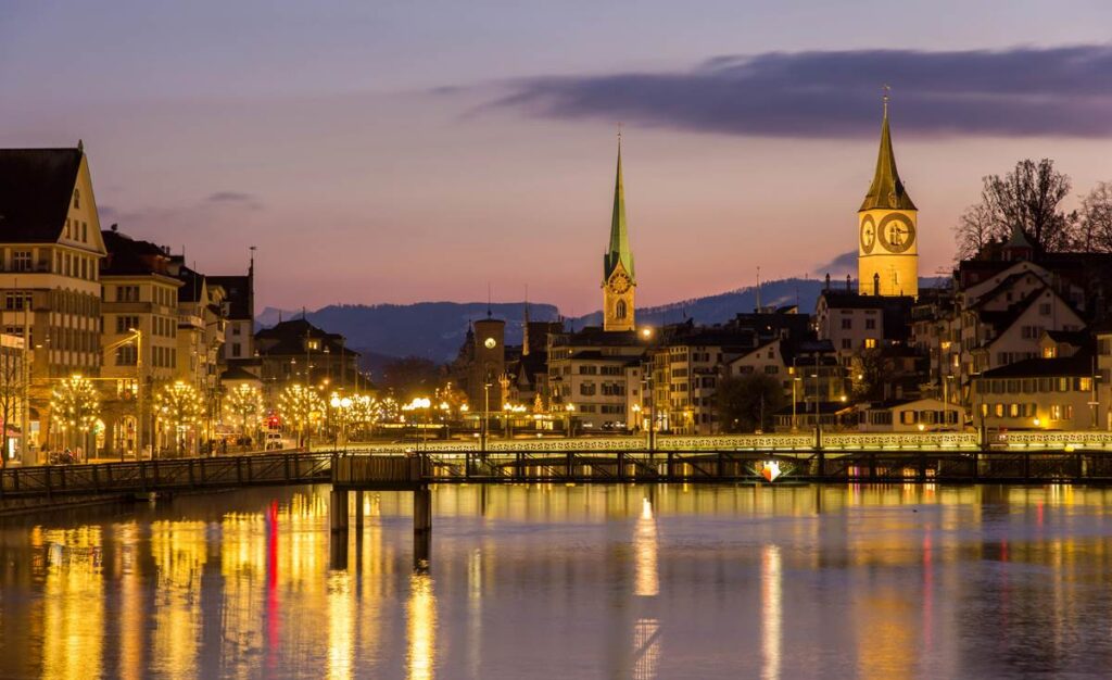 Night view of Zurich cityscape with illuminated buildings reflecting on the river under a twilight sky.
